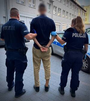 Policjant i policjantka prowadza zatrzymanego w kajdankach.