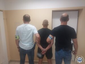 Dwaj funkcjonariusze trzymają zatrzymanego mężczyznę zakutego w kajdnaki.