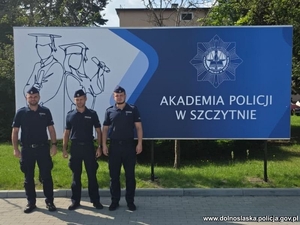 Trzej policjanci na tle tablicy z napisem Akademia Policji w Szczytnie.