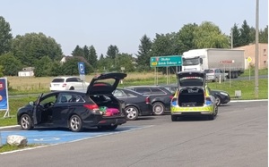 Zaparkowane samochody, jeden z nich ma otwarte drzwi od strony kierowcy i otwarty bagażnik. Z tyłu stoi radiowóz, którego bagażnik tez jest otwarty.
