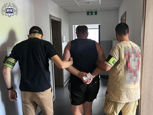 Dwaj funkcjonariusze prowadzą zatrzymanego mężczyznę zakutego w kajdanki.