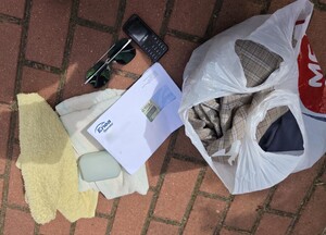 Na ziemi leżą przedmioty zabezpieczone przy zatrzymanym mężczyźnie: telefon komórkowy, okulary przeciwsłoneczne, koperta, foliowa reklamówka z odzieżą.
