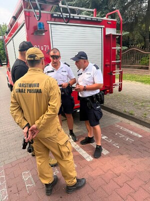 Dwaj policjanci i dwaj strażacy stoją przy wozie strażackim.