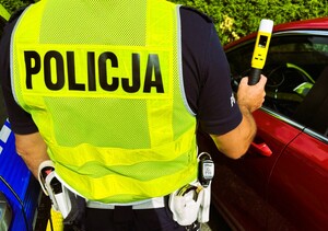 Widziany od tyłu funkcjonariusz ruchu drogowego z alkotesterem w prawej dłoni stoi przy samochodzie osobowym.