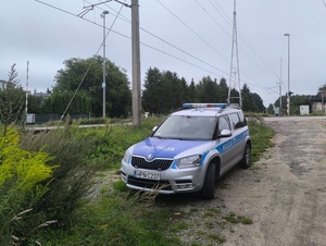 Radiowóz policyjny stoi na poboczu blisko słupa linii trakcji kolejowej.