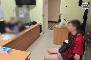 Siedzący przy biurku policjant przesłuchuje zatrzymanego siedzącego na krześle naprzeciw.