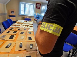Na blacie stołu leżą zabezpieczone przez policjantów telefony komórkowe i smartfony. Obok stoi policjant robiący notatki.
