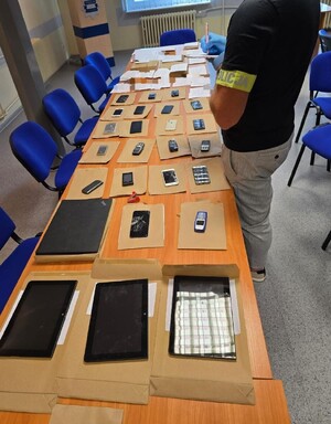Na blacie stołu leżą zabezpieczone przez policjantów telefony komórkowe i smartfony. Obok stoi policjant robiący notatki.