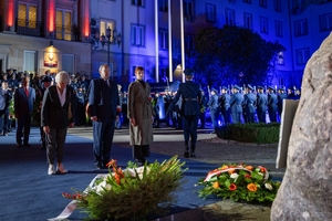 Trzy osoby oddają hołd poległym policjantom przed obeliskiem na terenie Komendy Głównej Policji. W tle uczestnicy uroczystości stojący przed budynkiem KGP.
