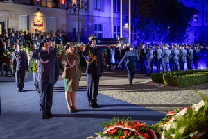 Delegacje oddają hołd poległym policjantom przed obeliskiem na terenie Komendy Głównej Policji. W tle uczestnicy uroczystości stojący przed budynkiem KGP.