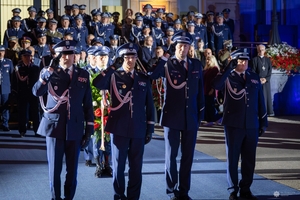 Delegacja złożona z czterech policjantów oddających hołd poległym policjantom przed obeliskiem na terenie Komendy Głównej Policji. W tle uczestnicy uroczystości stojący przed budynkiem KGP.