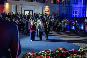 Trzy kobiety oddają hołd poległym policjantom przed obeliskiem na terenie Komendy Głównej Policji. W tle uczestnicy uroczystości stojący przed budynkiem KGP.