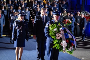 Delegacja z wieńcem kwiatów przed obeliskiem na terenie Komendy Głównej Policji. W tle uczestnicy uroczystości stojący przed budynkiem KGP.