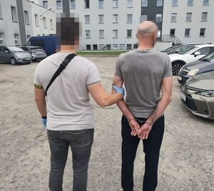 Policjant trzyma za ramię zatrzymanego mężczyznę zakutego w kajdanki.