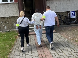 Policjantka i policjant prowadzą w kierunku wejścia do budynku zatrzymanego mężczyznę.