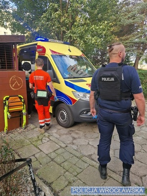 Odwrócony tyłem policjant, w głębi karetka pogotowia i ratownik medyczny.