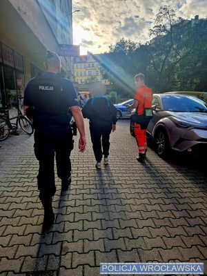 Ratownik medyczny i policjant idą z kobietą chodnikiem.