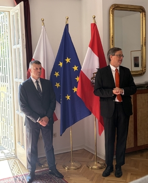 Dwaj mężczyźni stoją w pomieszczeniu ambasady na tle flag Polski, Austrii i Unii Europejskiej.