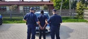 Dwaj funkcjonariusze  z zatrzymanym mężczyzną zakutym w kajdanki stoją przed radiowozem.