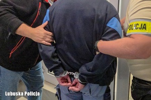 Dwaj funkcjonariusze trzymają zatrzymanego mężczyznę zakutego w kajdanki.