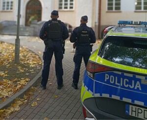 Dwaj policjanci przed budynkiem szkoły, obok nich stoi zaparkowany radiowóz.