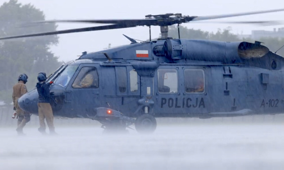 Śmigłowiec Black Hawk w deszczu na płycie lotniska, przed jego dziobem po obu stronach stoją policyjni lotnicy.