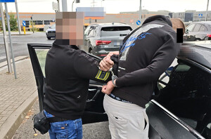 Policjant trzyma zatrzymanego mężczyznę zakutego w kajdanki przy radiowozie.