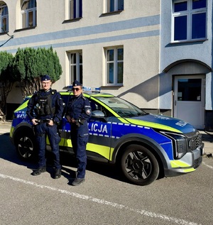 Policjanci pozują do zdjęcia przed radiowozem zaparkowanym przed budynkiem Policji.