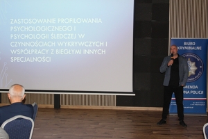 Mężczyzna stoi na tle bannera z logiem Biura Kryminalnego Komendy Głównej Policji. Po lewej stronie na ekranie wyświetlana jest prezentacja.