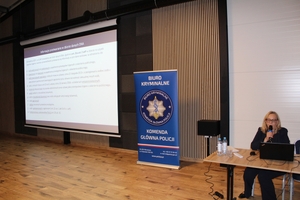 Kobieta siedzi przy stole, przed sobą ma otwarty laptop. Obok niej puste krzesło. Po lewej stronie banner z logiem Biura Kryminalnego Komendy Głównej Policji. Dalej na ekranie wyświetlana jest prezentacja.
