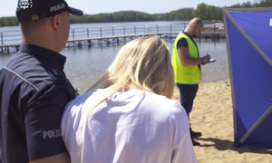 Na plaży kobieta z pochyloną głową stoi obok umundurowanego policjanta, w tle policjant w kamizelce odblaskowej robi notatki.