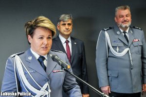 Komendant Wojewódzki Policji przemawia do zgromadzonych -  w tle policjant i inny mężczyzna.