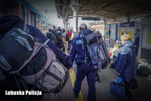Policjant pomagający nieść plecak kobiecie na peronie kolejowym.