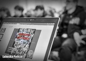 Czarno białe zdjęcie przedstawiające laptop, na wyświetlaczu którego widnieje prezentacja o Zbrodni Katyńskiej, a w tle uczestnicy spotkania.