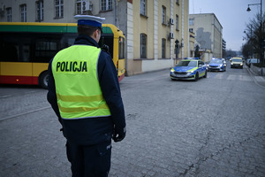 Policjant Ruchu Drogowego w kamizelce odblaskowej z napisem Policji w tle radiowóz.