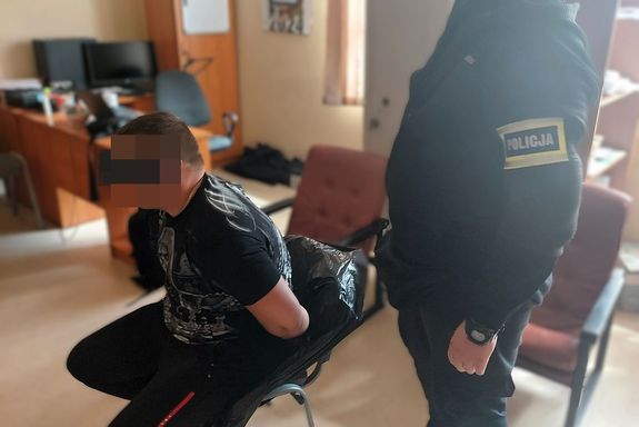 Zatrzymany siedzi na krześle w pokoju ma kajdanki założone na ręce trzymane z tyłu, obok stoi policjant.