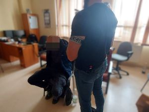 Zatrzymany siedzi na krześle, ma kajdanki założone na ręce trzymane z tyłu, obok stoi policjant.