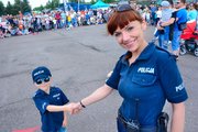 uśmiechnięta policjantka z chłopcem, który ciągnie ją za reke, ubrany jest w mini mundur policyjny ,w tle zgromadzeni przed pokazem goście festynu :dzieci i dorosli