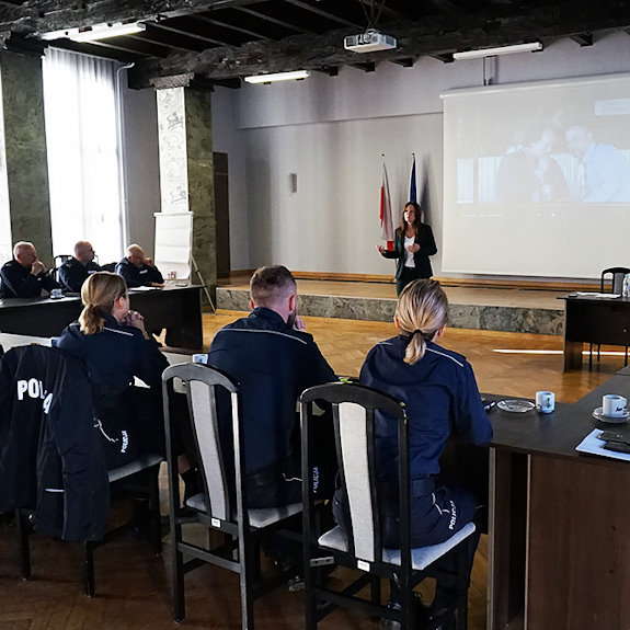 Kobieta przemawia do siedzących za stołami policjantów, na ekranie za nią wyświetlony kadr z filmu.