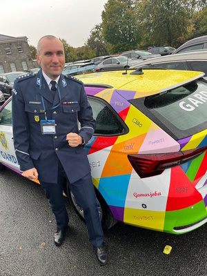 Funkcjonariusz polskiej Policji w umundurowaniu służbowym stoi obok radiowozu policji irlandzkiej. Samochód ozdobiony jest kolorową grafiką z napisami „dzień dobry” w różnych językach, symbolizującą różnorodność kulturową i otwartość irlandzkiej Policji.