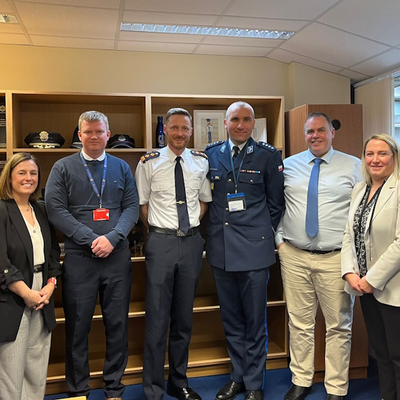 Grupa uczestników spotkania w siedzibie policji irlandzkiej w Dublinie pozuje wspólnie do zdjęcia. Wśród nich znajduje się funkcjonariusz polskiej Policji w umundurowaniu służbowym oraz przedstawiciele irlandzkiej Policji i Międzynarodowego Stowarzyszenia Policji  IPA.