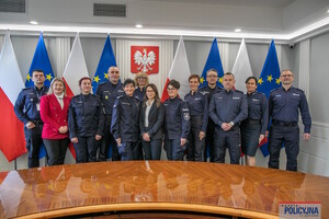 Grupa osób stojących na tle flag Polski i Unii Europejskiej. Są to osoby uczestniczące w spotkaniu- kobiety i mężczyźni, policjanci i pracownicy Policji.