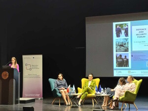 Kobieta za mównicą, cztery panelistki siedzą w fotelach, w tle na ekranie plakat konferencji.
