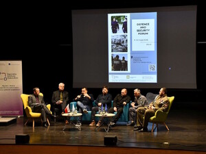Siedmiu panelistów siedzi w fotelach, jeden mówi do mikrofony, w tle na ekranie plakat konferencji.