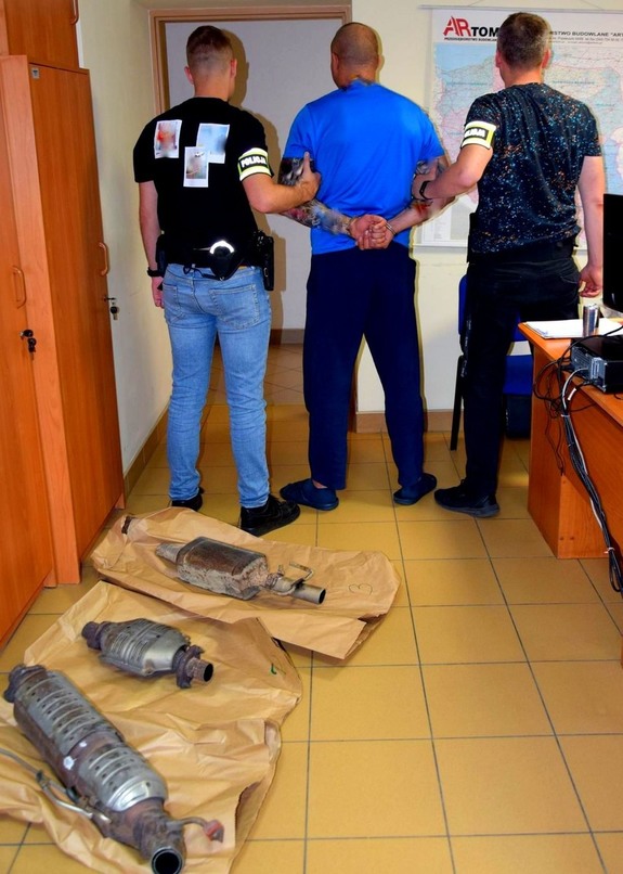 Mężczyzna stojący tyłem z kajdankami na rękach po obu stronach policjanci z opaską na ręce policja. Na podłodze leżą 3 katalizatory.
