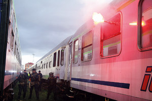 Grupa policjantów z bronią gotową do strzału idzie pomiędzy dwoma składami pociągów. Przez okno jednego z wagonów wystawiona zapalona raca.