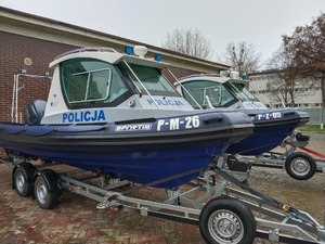 Dwie policyjne łodzie na przyczepach do transportu. Na burcie pierwszej napis P-M-26 na drugiej P-Z-05.