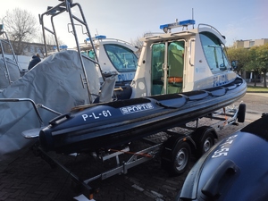 Pojedyncza niebiesko-biała policyjna łódź patrolowa sfotografowana z tylnej bocznej strony, stojąca na przyczepie do transportowania łodzi, z widocznymi elementami znajdującymi się w tylnej części łodzi i tyłem oraz wejściem do kabiny ze sterem