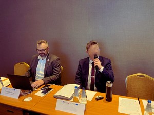 Na zdjęciu przedstawieni zostali siedzący przy biurku przed ciemnoszarą ścianą przedstawiciele CBŚP, z których jeden patrzy w komputer, a drugi trzyma mikrofon.