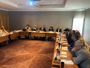 Zdjęcie przedstawia siedzących przy trzech prostokątnych stołach ułożonych w podkowę uczestników spotkania.